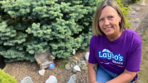 BBC / Simon Spark Niki Hall. She has short, light brown hair and is wearing a purple T-shirt with the wording Laybo's Legacy - Let's change things together.