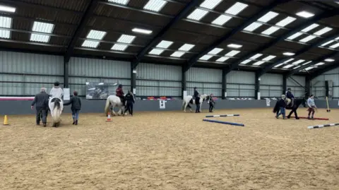 A view of the horse riding school. There are four horses, with four riders on them. The ground is light brown and there are blue and white tubes on the ground. There are people stood either side of the horses, leading them around the school.