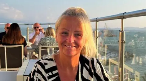 Suffolk Police A photo of Anita Rose smiling directly into the camera. She has blonde hair, which is tied up, and is wearing a black and white patterned top. She appears to be sitting at a table in a restaurant on a high-rise terrace. Other diners can be seen in the background.