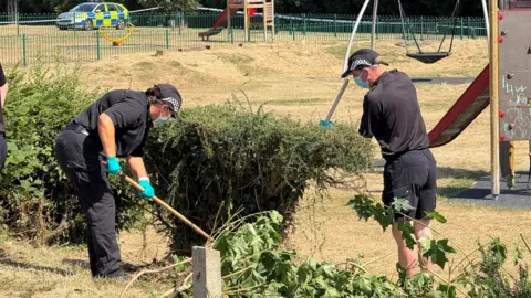 Steve Hubbard/BBC Two police officers dressed in black uniforms appear to be searching a green bush which is beside a play area. They wear blue gloves and face masks. A marked yellow and blue police car is in the background.