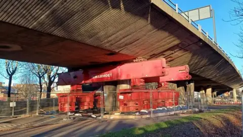 A large, red prop supporting the concrete bridge. The metal prope surrounds one of the existing suports to hold the bridge up.