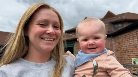 Leah Morrison, who has long blonde hair and is wearing a grey coloured sweater, is holding her son Niall, who is smiling at the camera and is wearing a blue bib and a very light pink coloured jumpsuit.
