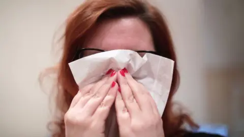 A woman with bright reddish pink nail varnish sneezes into a tissue. She has black framed glasses and auburn hair.