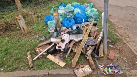Harry Machin A pile of rubbish by the side of a road. There are a number of vapes in a metal basket, the rubbish includes a water can, toys, wood, lots of green and blue waste bags, and other items. They are sitting on a patch of grass. 