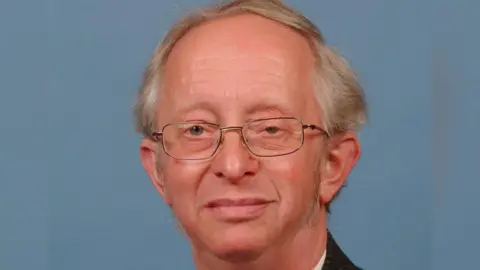 A close-up showing the head of a man wearing silver wire-framed glasses. He has grey hair in a side parting and the collar of a dark suit and white shirt can just be seen. The image is against a blue background.