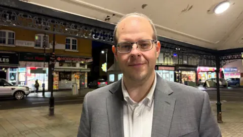 Martin Heath/BBC Jonathan Ash-Edwards with short grey hair and glasses, wearing a grey jacket and white shirt. He is standing under a covered pavement to which Christmas decorations have been attached and there are shops in the background.