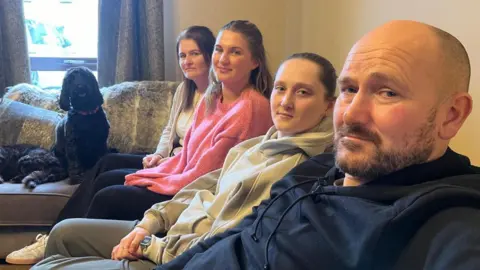 Lachlan is sitting on a sofa with his two daughters and his wife Leeann. The family's dogs have couch all to themselves.