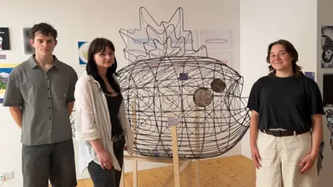 On the left there is a male student wearing a grey shirt and black shorts, a female student wearing a a white overshirt and black jeans, then the metal fish sculpture in the middle with the final female student on the right wearing a black top and white shorts.