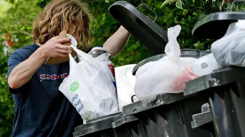 A man, whose long wavy blond hair obscures his face, empties waste in a white plastic carrier bag into a grey wheelie bin.