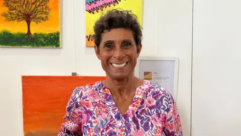 Fatima Whitbread looks at the camera and smiles. Behind her there is a white wall with paintings on it, including one of a tree in front of an orange sky.