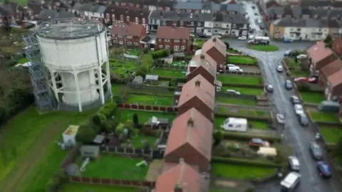 A white water tower in a field surrounded by several houses. 