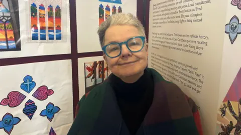 A woman with short grey hair and blue glasses stands smiling in front of boards with information and designs of the new window.