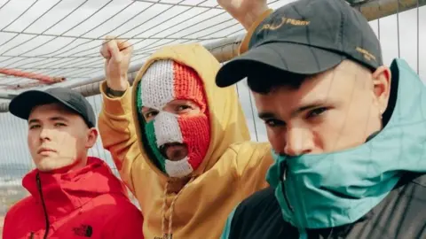 Shows three members of Kneecap, two wearing baseball caps and one with a balaclava in the colours of the Irish tricolour