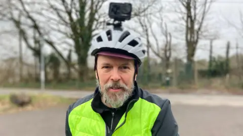 Guy Buckland wearing a high-vis jacket and a white helmet. His helmet has a camera attached to the top of it. He is standing outside on a cloudy winter day.