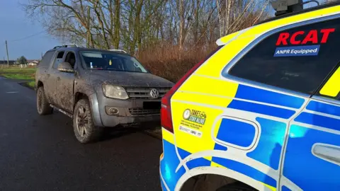 A four-by-four Volkswagen vehicle covered in mud parked on the side of the road with a police car in front of it.