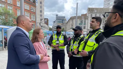Gary Godden has short hair and is wearing a suit. Claire Ward has long brown hair and is wearing a pink overcoat. They are stood talking to security officers in high visibility uniform. 