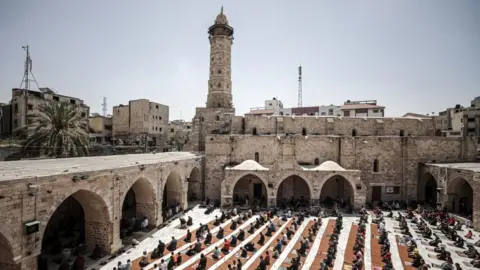 The World Monuments Fund / Getty A stone coloured mosque in the shape of a square, there are arches surrounding the square. In the centre of the mosque is rows of people praying on mats. In the left hand corner of the mosque is a stone coloured tower, the background is also filled with various stone buildings.