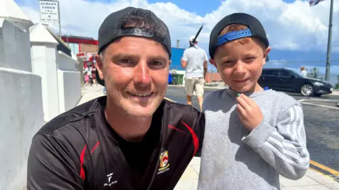 Simon Patterson and his son Sam are both smiling as Simon, on the left of the image, kneels down and son Sam stands. Simon is wearing a black baseball cap and black football team shirt. Sam has one hand up to his mouth and is wearing a grey sweatshirt and baseball cap. Both Simon and sam have their caps turned backwards.