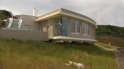 A modern, single-story building with a curved facade and large windows is situated on a grassy bank above a concrete sea wall. The building has a flat roof and features an outdoor terrace with glass railings. The sky is overcast.