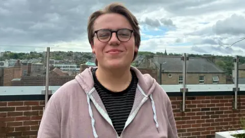 A young man stands in front a low red-brick wall, with a view of Barnsley skyline behind him. He has short dark blonde hair and glasses, and wears a black t-shirt with white stripes and a light pink hoodie.