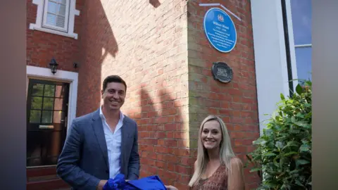 Trafford Council A man with short dark hair and wearing a white shirt and grey jacket and a woman with long blonde hair and wearing a brown patterned top are stood smiling in front of a red brick house where a blue plaque has been unveiled.