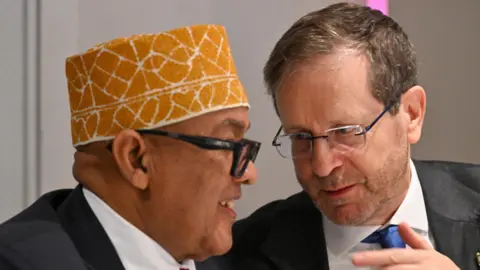 Israeli president’s office / X The Somaliland president, Abdirahman Mohamed Abdullahi, wearing a black suit, white shirt and red tie with traditional hat is talking to the President of Israel, Isaac Herzog. Herzog is wearing a black suit, white shirt and blue tie. They both are wearing glasses.
