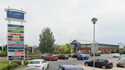 Google St John's Retail Park, Bedford, showing a large sign, a number of cars, buildings in the distance and a street lamp.