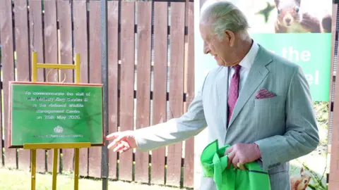 PA Media King Charles III, who wears a grey suit, points to a green plaque commemorating his visit to the National Wildlife Management Centre.