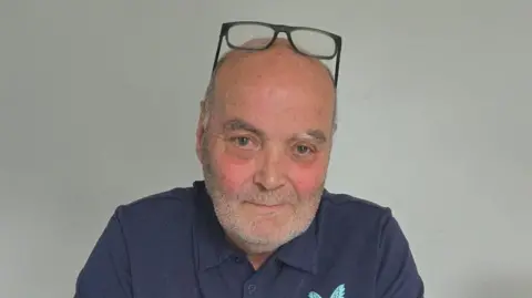 Dave Kelly Chairman Dave Kelly sits in front of a plain wall in a navy blue polo shirt.