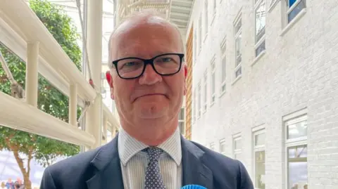 Vikki Irwin/BBC Matthew Hicks is standing in a bright atrium, with a tree in the background. He is wearing a blue suit, a white and blue striped shirt and blue patterned tie. He is wearing glasses and smiling at the camera.