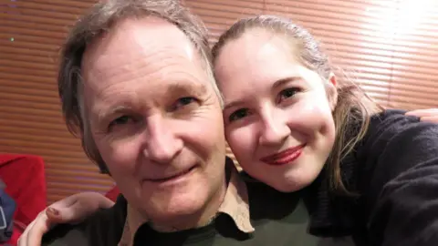 Family handout A man with grey hair and his adult daughter have their arms around each other and are looking into the camera and smiling. 