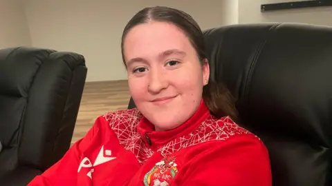 Paige, 17, smiles towards the camera wearing a red Wales FC zip up jacket. Has brown hair tied back. 