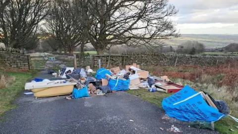 Litter Free Ilkley A load of trade waste including an old bath and several large blue bags filled with rubbish, dumped on Ilkley Moor. There is a stone wall, trees and fields and moors in the background. 
