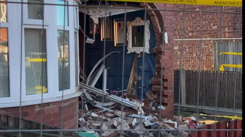 Close-up image of brick damage to the house's front entrance and interior. Paintings and a mirror still remain intact on the wall of the hallway.