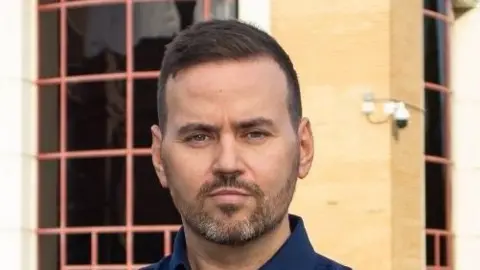 Basildon Council Gavin Callaghan smiles at the camera. He has short dark hair with a beard and wears a navy blue shirt. A yellow brick building can be seen behind him with large windows.