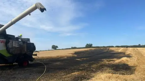 Cleveland Fire Brigade A combine harvester which has been burned in a fire sits in a large field on a sunny day. The harvested is black and there is a water hose leading up to the machine. The hay which is surrounding the machine is scorched black from a fire. 