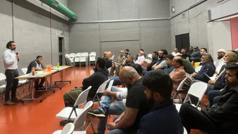 Aisha Iqbal/BBC A community hall with people seated on rows of chairs. A man stands at the front holding a microphone and addresses them.