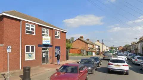 Google A narrow street with cars parked either side. There is a large red brick building to the left and houses further along the street.