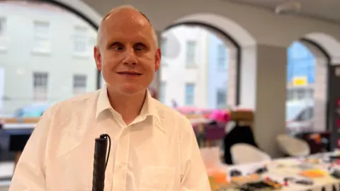 Dave smiles at the camera as he wears a white shitr and holds his walking cane. Behind him is a desk with lots of braille equipment on it.