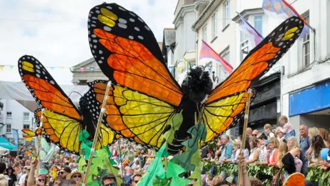 Penzance Council Large butterfly puppets with bright orange and black wings are carried above a dense crowd as a parade winds through a bunting‑filled town centre packed with onlookers.