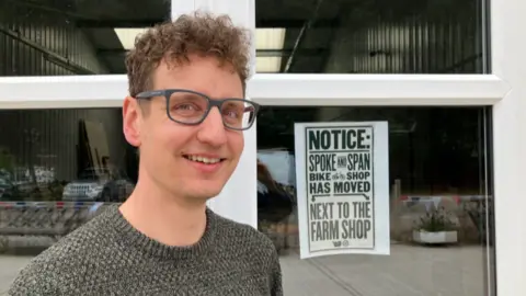 A man with curly hair and glasses, wearing a grey jump is stood in front of a window. A sign in the window reads Notice: Spoke and Span Bike Shop has moved