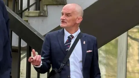 A man with a bald head and no teeth is mid-conversation as he walks down a stairway outside a court. He is wearing a navy blazer, a white shirt, a red, navy and white tie, and a black crossbody bag. He is holding a vape.
