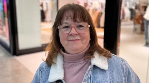 Sharon Chambers, a woman wearnig a pink jumper and a denim jacket. She has light brown hair and is standing indoors inside a shopping centre.