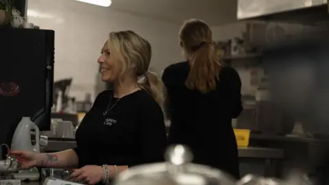 Kim is standing behind the counter of a café kitchen. She is wearing a dark T‑shirt with light‑coloured text on the chest. The background includes stainless‑steel surfaces, shelves with various containers, and kitchen equipment. In the foreground, out‑of‑focus objects such as a glass cake dome and small appliances partially frame the scene, giving the impression of a busy working environment.
