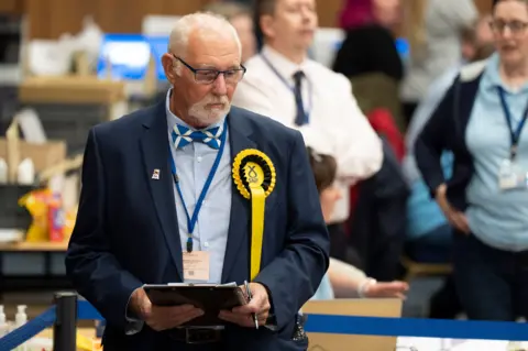 PA Media A serious looking SNP counter in Saltire bow-tie and blue suit stands with a clipboard watching the vote tally in a hall full of people.