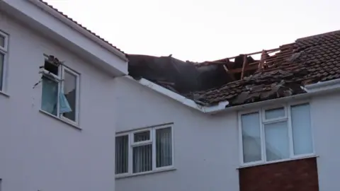 A view of the roof of the building showing part of it missing. The window on the left is also broken. The building is white. The sky is cloudy. 