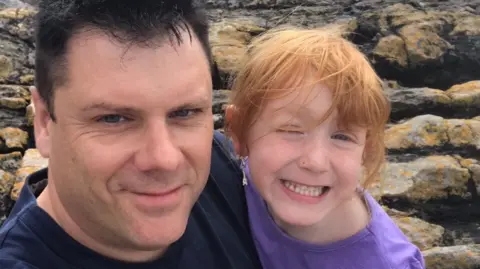 Andrew McQueen Andrew and his daughter Poppy are standing arm in arm by some rocks. They are both smiling at the camera.