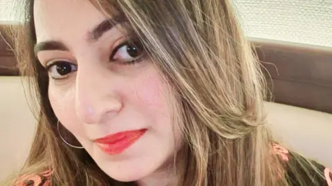 Yaditi Kava has long blonde hair and red lipstick. She is wearing a black and pink dress.