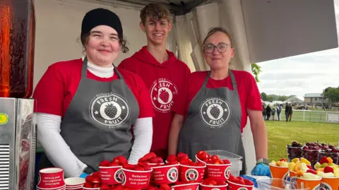 Three people wearing red behind a stall counter at the Bath and West Show smiling at the camera with small boxes of strawberries and other fruits sitting in front of them. On their aprons, there is a circular logo, with the words "fresh fruit" written above and below a picture of a strawberry.