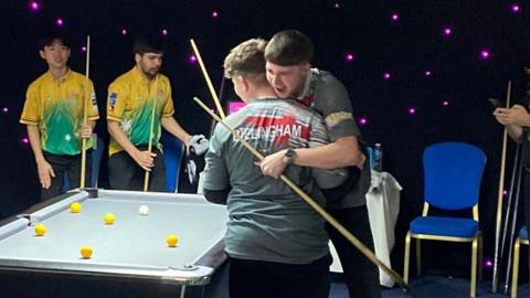 Two young players embrace beside a pool table. Both wear grey T-shirts with red flats on them. Both hold pool cues, one has light hair, the other dark. Beside them is a pool table with six balls on it, one white, five yellow. The table has a grey, cloth top. Behind the table are their two opponents, both holding cues, in yellow and green T-shirts
